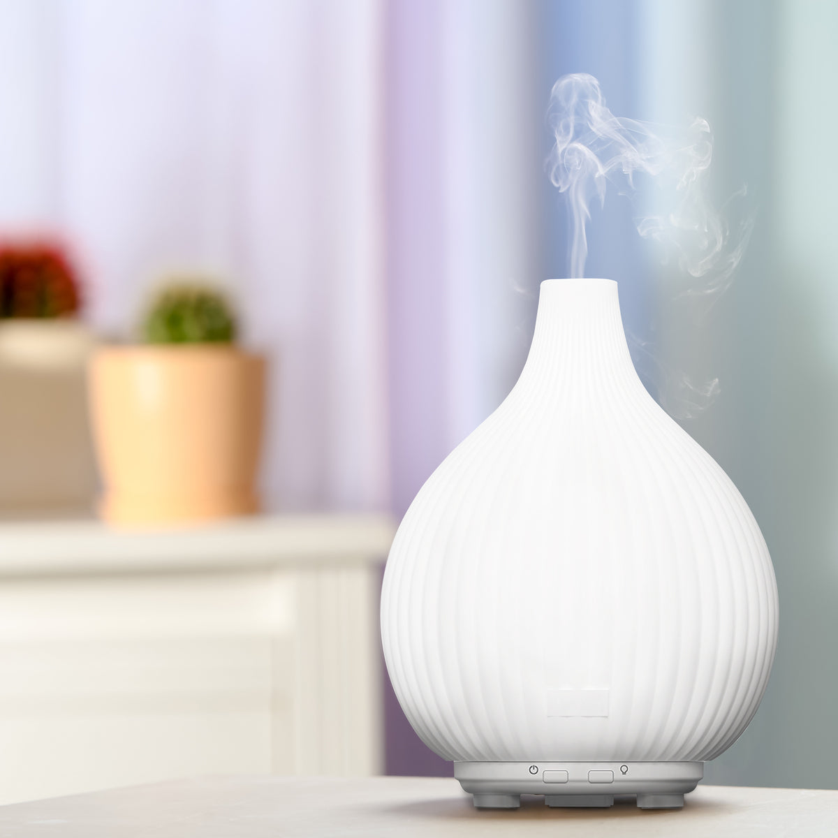 The Rio LYLA Aroma Diffuser, Humidifier & Night-Light sits on a table emitting vapor, creating a relaxing atmosphere. The white ribbed design stands out against a background of potted plants and soft pastel curtains.