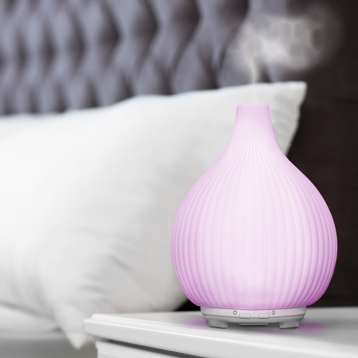 The LYLA Aroma Diffuser, Humidifier & Night-Light by Rio emits mist on a white bedside table, creating a relaxing atmosphere beside a bed with a white pillow and tufted dark headboard.