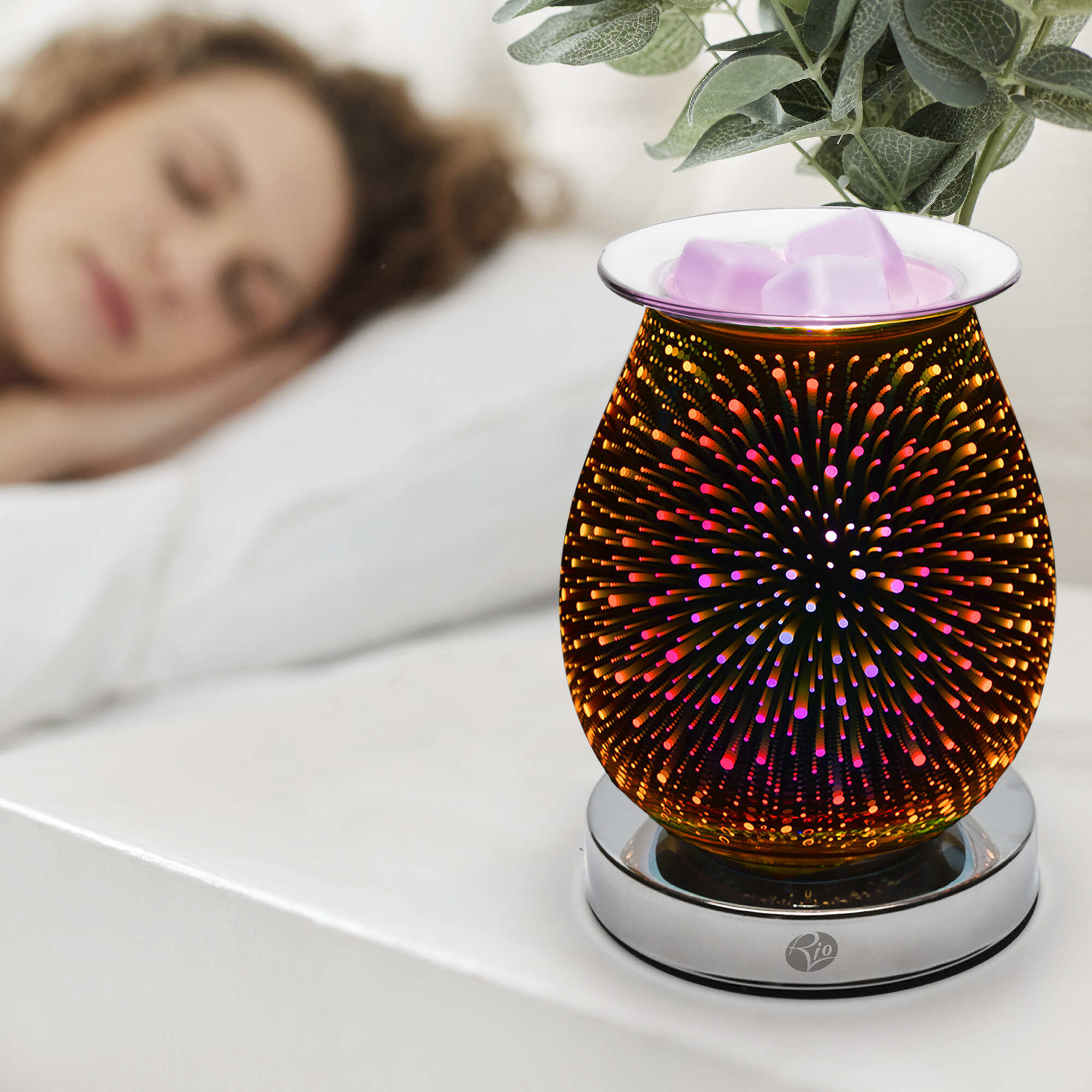 A woman sleeps in the background while the Rio Alura Wax Melt Burner & Aroma Diffuser Lamp, glowing with a colorful starburst pattern and melting pink wax cubes, sits on her bedside table beside a potted plant for a cozy touch.