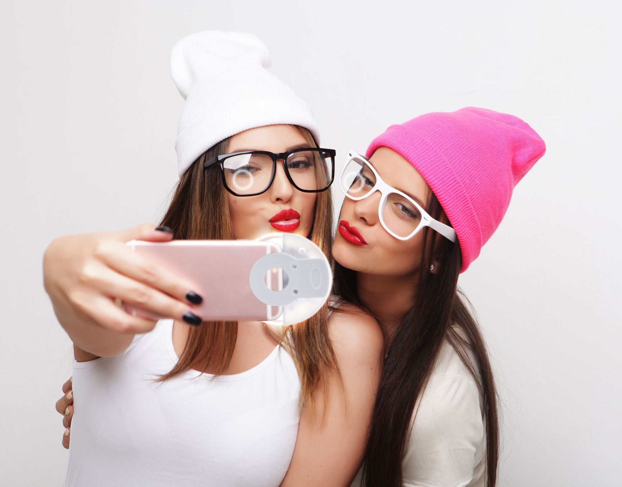 Two women in colorful beanies and glasses pout with red lipstick and smile for a selfie. One holds a smartphone fitted with the Rio Beauty Glow Smartphone Ring Light, capturing their fun moment against a plain white background.