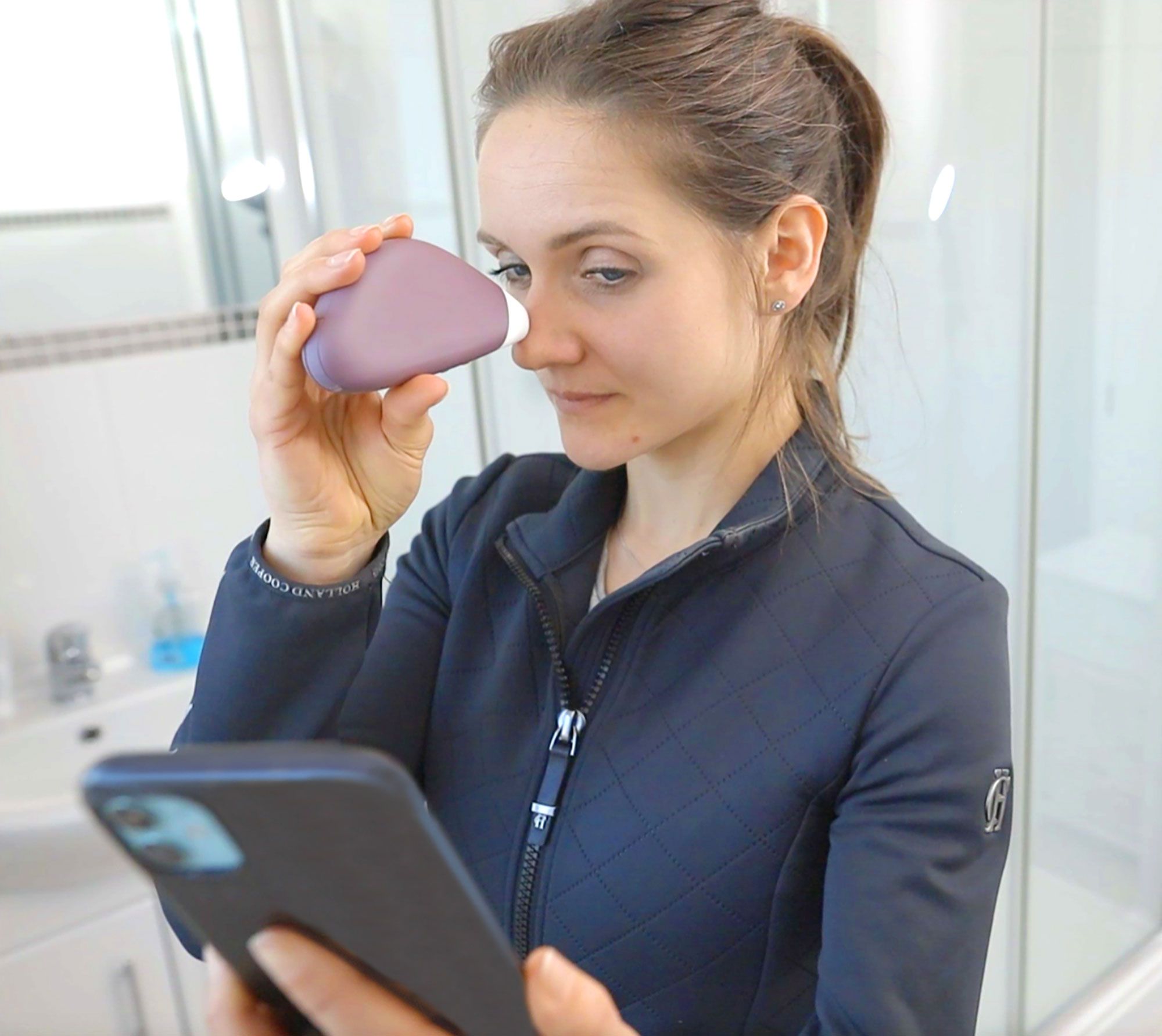 lady in bathroom using pore perfector wireless purple device on nose while holding and looking at smartphone