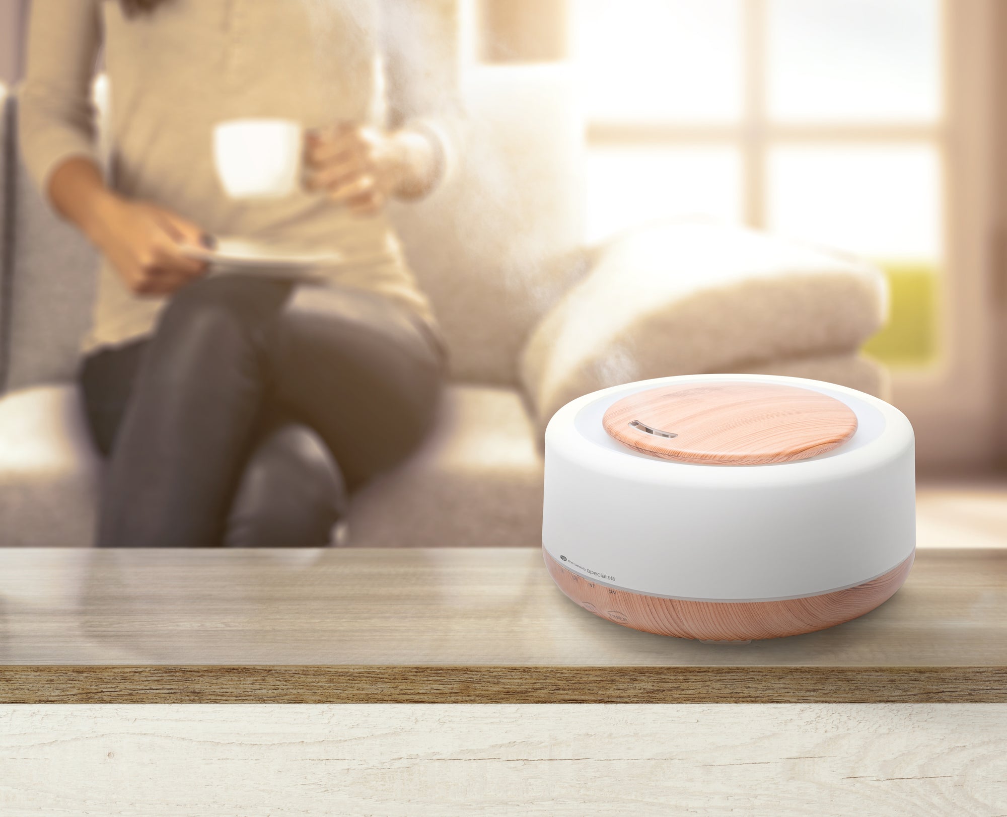 The Rio ALORA Aroma Diffuser, Humidifier & Night-Light in white and wood style emits mist on a table, as a person relaxes with a cup on a sunlit couch in the background.